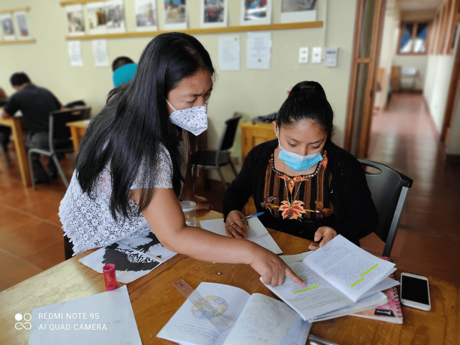 Mayan lady points to a book over a table where another girl is sat